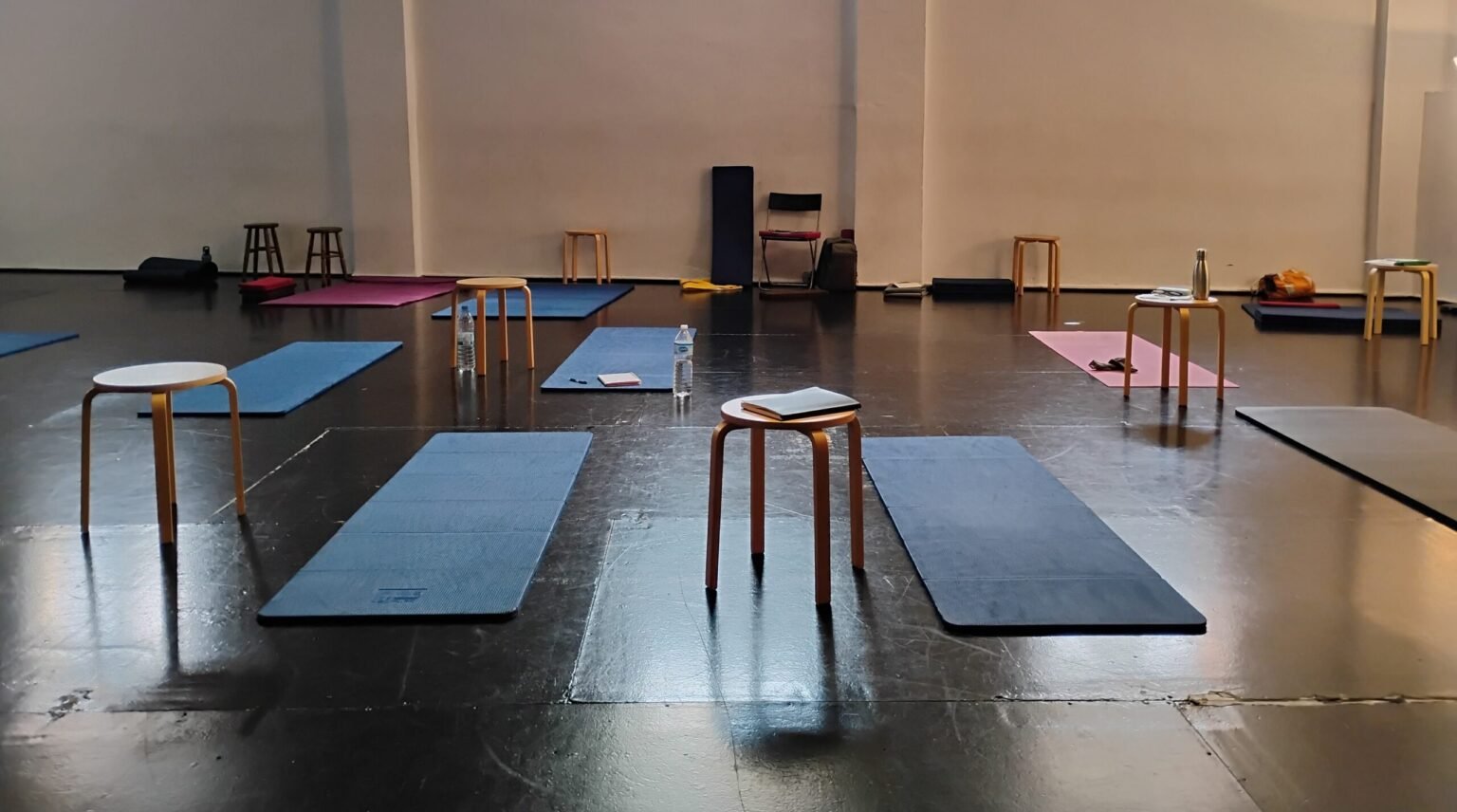 Un estudio amplio con suelo oscuro y brillante, organizado con esterillas de yoga de varios colores y taburetes de madera para una sesión grupal.