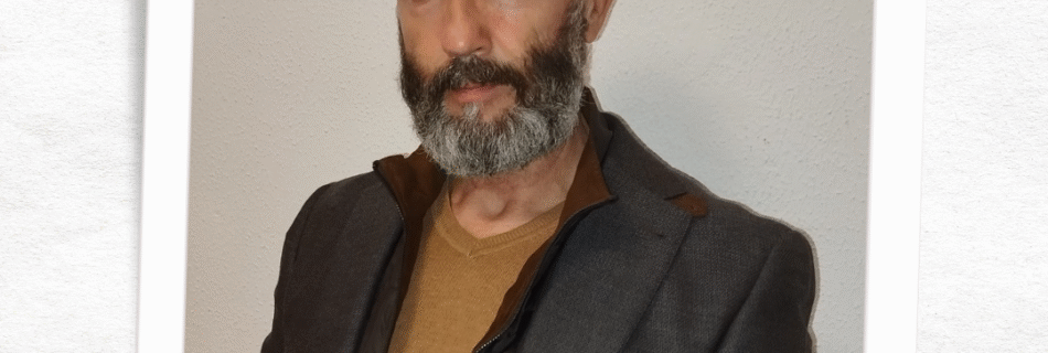 Retrato formal de David Sánchez, guía espiritual con más de 15 años de experiencia en Tarot y Numerología. Se presenta en un encuadre tipo fotografía polaroid con diseño floral minimalista, viste una chaqueta gris oscura sobre un jersey color mostaza. Tiene barba gris y es calvo. Su nombre 'David' aparece escrito debajo de su foto.